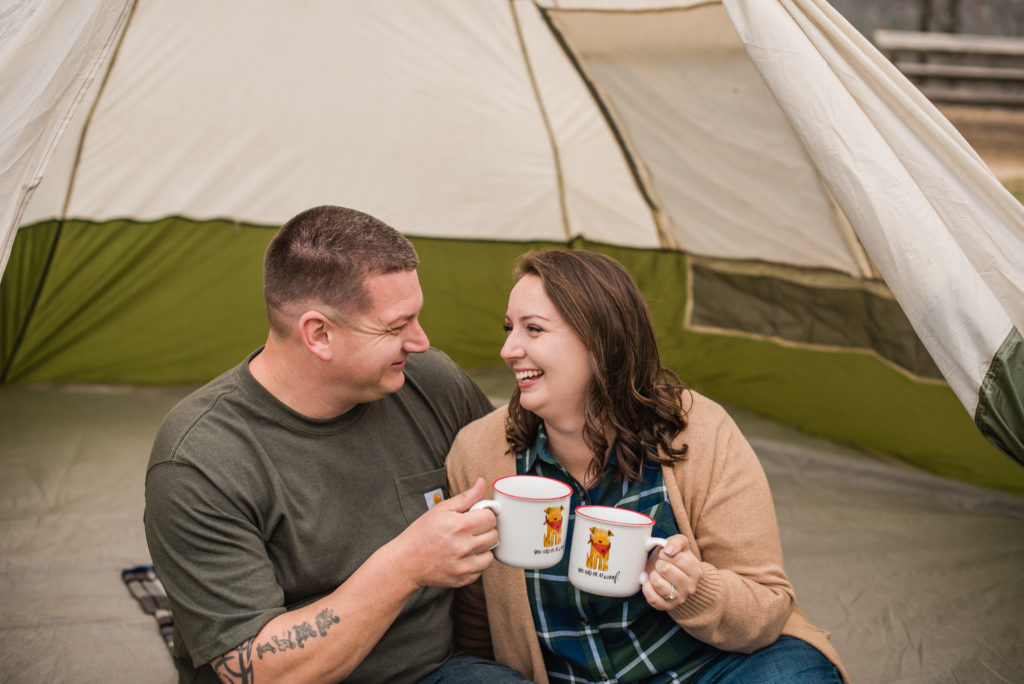 Couple wearing green toasting with coffee mugs in tent at their engagement session at FD Roosevelt State Park